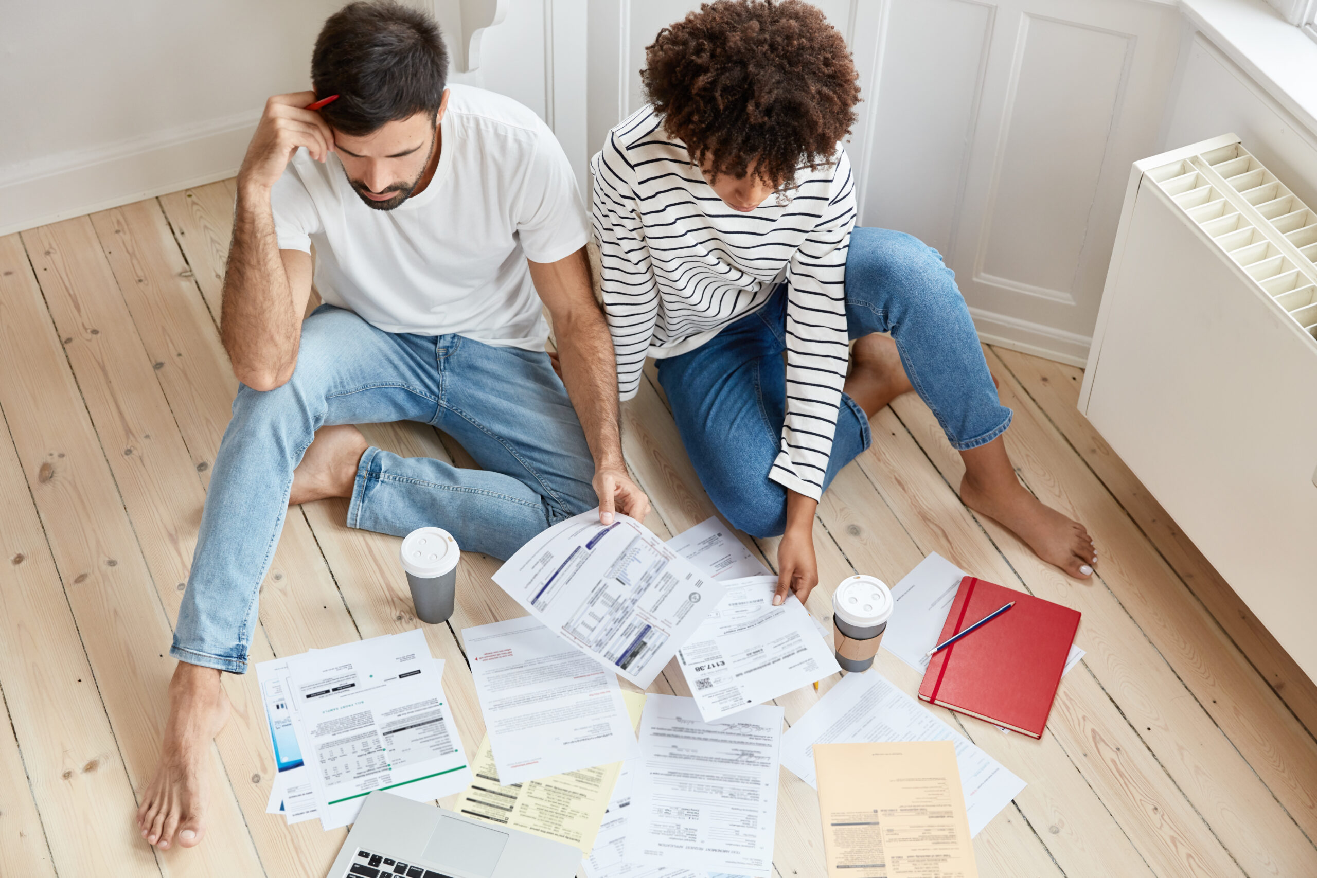 Top view of mutiethnic business partners works in domestic atmosphere, sit on floor and discuss financial chart, study home purchase contract together, drink takeaway coffee, involved in paper work